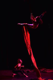 Photo of a performer in the air suspended by silks and another performer on stage looking up.