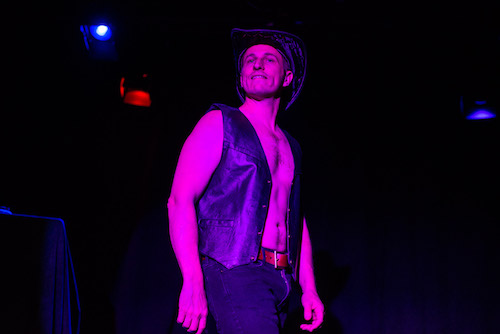 photo of DK Reinemer in Becoming Magic Mikeby Andrew Alexander - a shirtless white man in a leather vest and cowboy hat looks at the audience under purple stage lighting. 