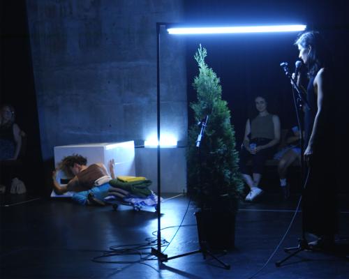 Person lying on dark stage touching white cube at their head, rest of cast of Bonjour Hi out of focus on sidelines