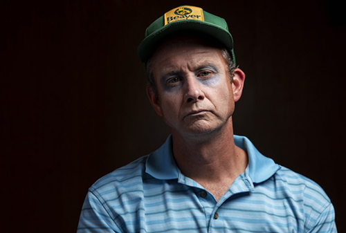 Man wearing pale make-up, a green ball cap, and a blue striped polo shirt stares at the camera, Footnote Number 12, SummerWorks