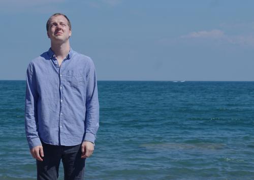 Man in jeans and blue shirt standing in front of ocean from Don’t Look Down Theatre Co’s. excellent The Ends of The Earth at Alumnae Theatre
