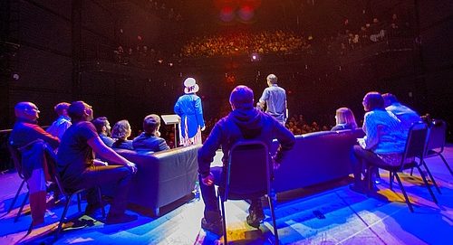 view from behind of several people sitting on a stage we see audience in background, from a performance of Your Hood's a Joke in Toronto