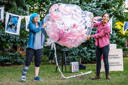 Photo of Kate Lushington and Natasha Greenblatt in an outside setting in a park, holding a clear bag filled with pink balloons between them in Apocalypse Play 